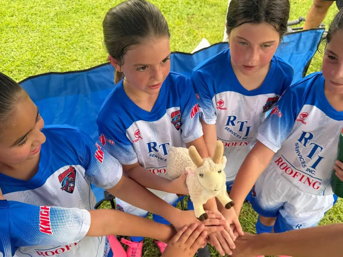 Girls youth soccer team in RTI Roofing jerseys stacking hands in a huddle around a stuffed goat mascot